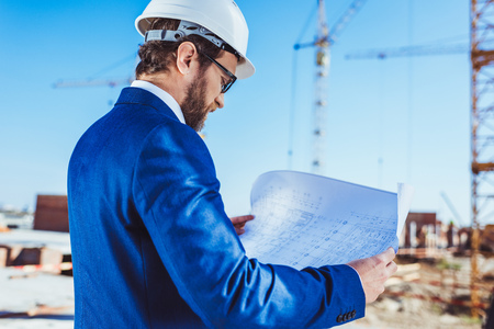 Businessman in hardhat and suit examining building plans at construction site