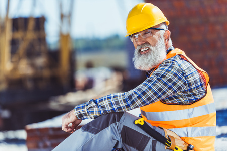 Bearded worker in reflective vest and hardhat sitting at construction site and looking at cameraの写真素材