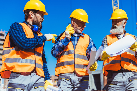Three workers in reflective vests and hardhats walking together and discussing building plans の写真素材