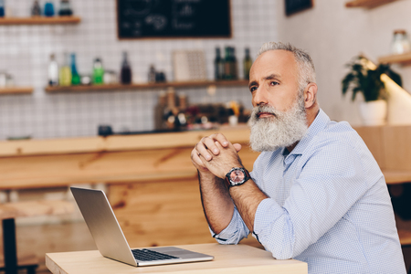 side view of thoughtful senior man sitting at table with laptop and looking away in cafeの写真素材