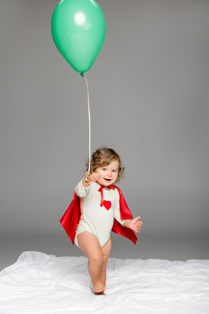 happy toddler in superhero cape holding balloon isolated on greyの写真素材