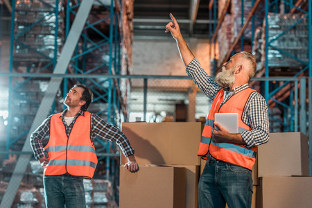 warehouse workers in vests holding digital tablet and pointing up with fingerの写真素材