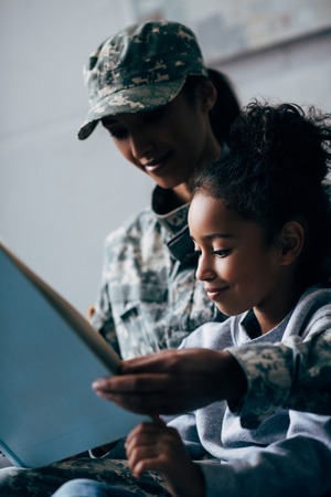 african american daughter and female soldier in military uniform reading book together at homeの写真素材
