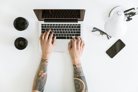 Top view of Workspace with male hands typing on laptop placed on white tabletop with camera lenses, glasses and smartphoneの写真素材
