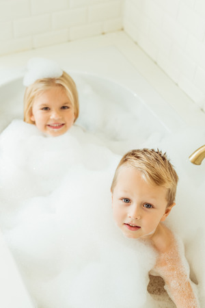adorable little kids smiling at camera while sitting together in bathtub with foamの写真素材