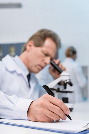 Concentrated lab technician taking notes while doing microscope sample analysisの写真素材