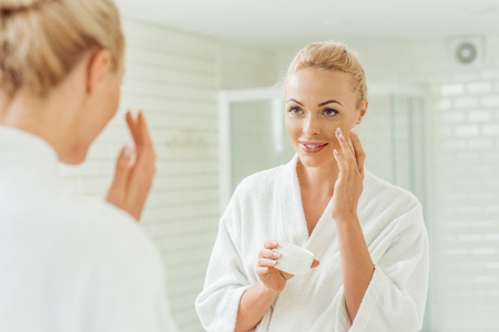 beautiful smiling young woman in bathrobe applying face creamの写真素材