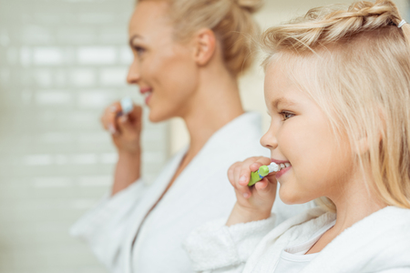 side view of happy mother and daughter in bathrobes brushing teeth togetherの写真素材