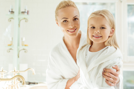 beautiful happy blonde mother and daughter in bathrobes smiling at camera  の写真素材