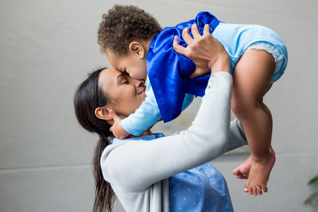 happy african american mother holding little son in superhero costume at homeの写真素材