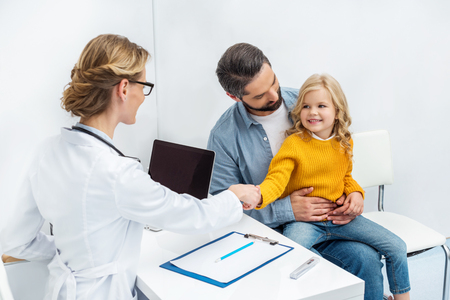 female doctor shaking hand of little girl while she sitting on fathers kneesの写真素材