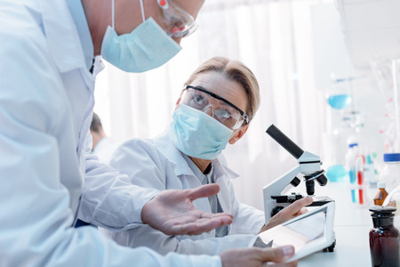 Doctors in protective glasses and masks working with microscope and discussing workの写真素材