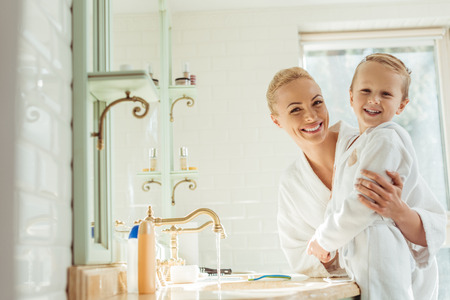 beautiful happy mother in son wearing bathrobes and smiling at camera in bathroom の写真素材