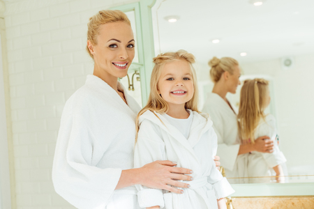 adorable happy mother and daughter in bathrobes smiling at camera in bathroomの写真素材