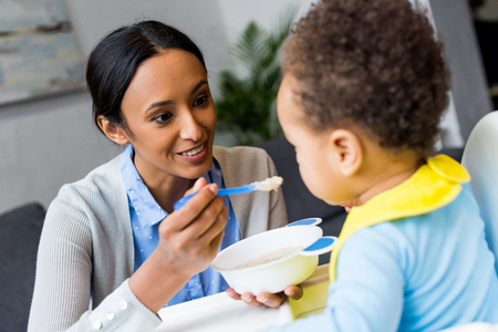 smiling african american mother feeding little son with porridge at homeの写真素材