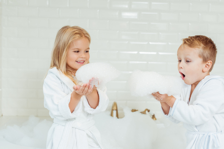 adorable little kids in bathrobes playing with foam in bathroomの写真素材
