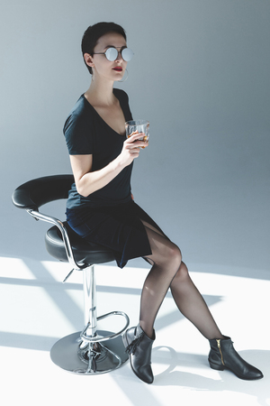 elegant young woman sitting on bar stool and drinking whiskey on greyの写真素材