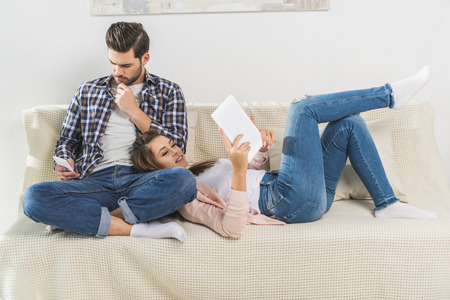 Young attractive couple using smartphone and digital tablet while sitting on sofaの写真素材