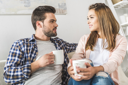 young attractive couple having coffee and sitting on couch at homeの写真素材