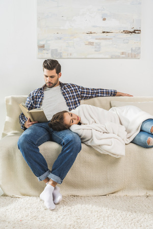 Young man sitting on couch and reading a book, while his girlfriend is napping on his knee の写真素材