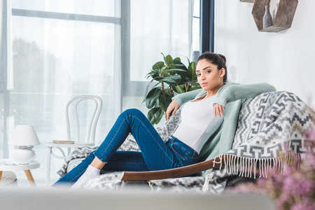 beautiful young woman sitting on couch and looking at camera at homeの写真素材