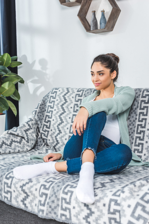 beautiful smiling young woman sitting on couch and looking away at home の写真素材