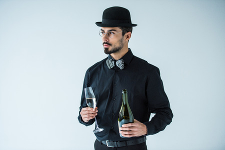 portrait of fashionable man in hat holding bottle and glass of champagne isolated on greyの写真素材