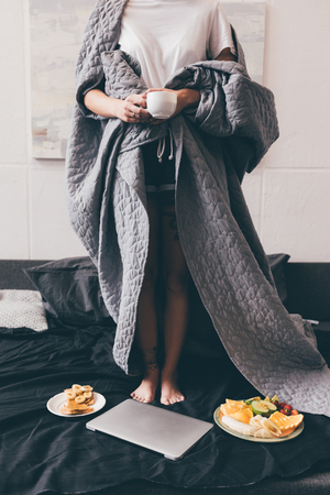 partial view of woman covered in blanket holding cup of coffee while standing on bed at homeの写真素材