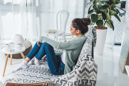 side view of pensive brunette girl sitting on couch and looking away at home の写真素材