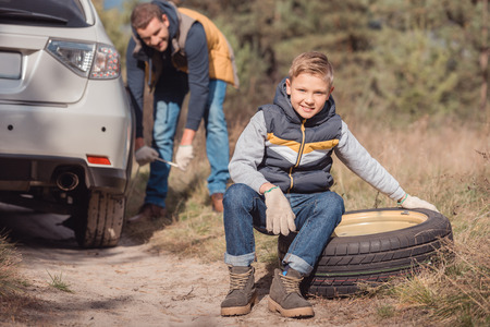 happy little son sitting on tire while father changing car wheelの写真素材