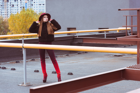 Attractive woman in burgundy hat and autumn outfit posing on a building roofの写真素材