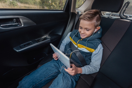 happy little boy in earphones using digital tablet while sitting in carの写真素材