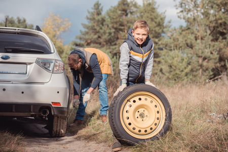 cheerful little boy smiling at camera while changing car wheel with father in forestの写真素材