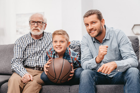 Little boy on couch with grandfather and father, cheering for a basketball game and holding a basketball ballの写真素材