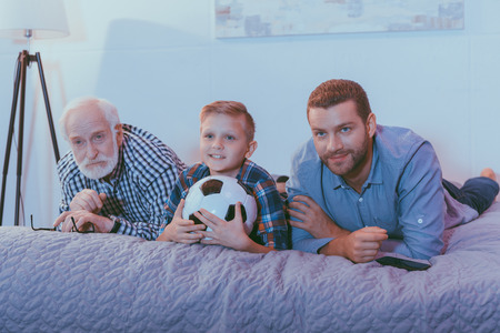 Little boy holding soccer ball, his father and grandfather lying on bed together and watching football gameの写真素材