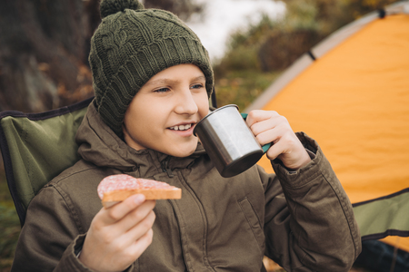 Preteen boy drinking tea and eating sandwich with sausage at the camping placeの写真素材