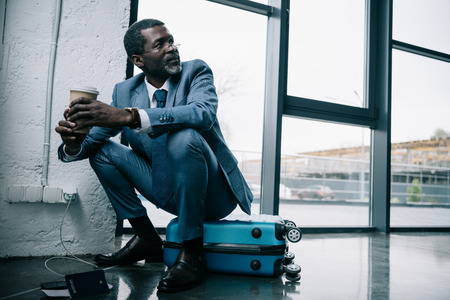 Middle aged African american businessman sitting on a suitcase at the airport and holding coffeeの写真素材