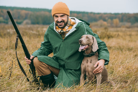 Handsome man squatting with a dog and gun and looking at cameraの写真素材