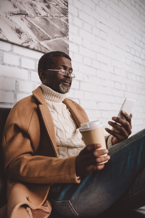 Middle aged African american man holding coffee in paper cup and looking at smartphoneの写真素材