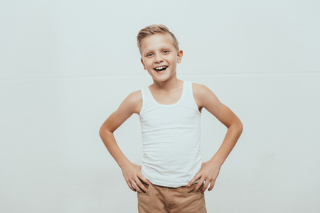 Young smiling boy in white tank top standing with hands on hips and looking at camera, isolated on whiteの写真素材