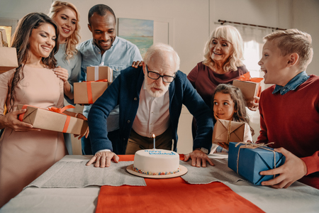 senior man blowing candle on birthday cake presented by large familyの写真素材