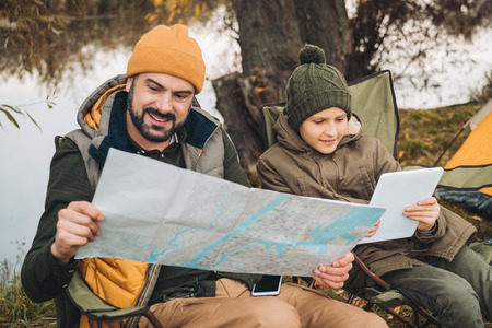 Father looking at map and son looking at tablet sitting on camp-chairsの写真素材