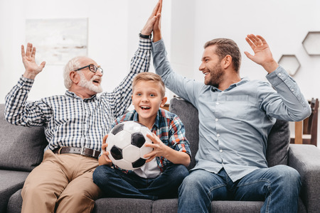 Little boy sitting on couch in living room and holding soccer ball, while his father and grandfather are high-fiving each otherの写真素材
