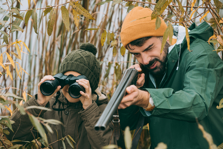 Father aiming at an animal with a gun, son looking through binocularsの写真素材