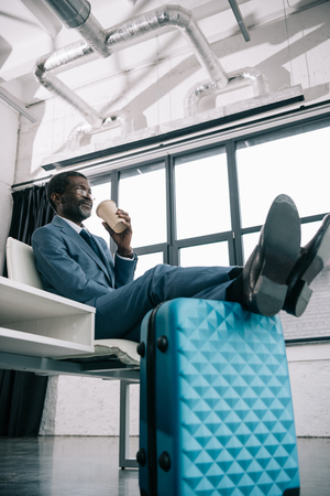 Middle aged African american businessman waiting at airport putting legs on suitcase and drinking coffeeの写真素材