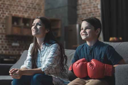 Little boy in boxing gloves sitting on couch with his mother and watching a boxing matchの写真素材