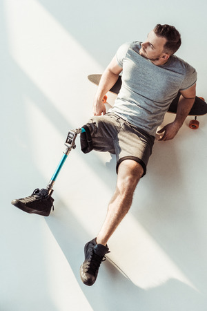 overhead view of pensive man with leg prosthesis resting on skateboarding isolated on whiteの写真素材