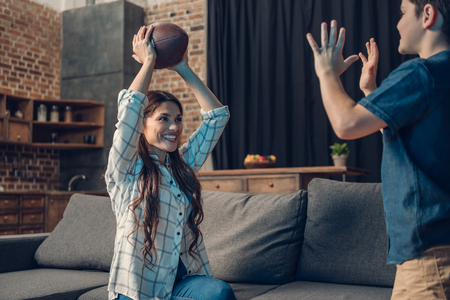 Little son and his beautiful mother playing with a rugby ball while sitting on couch in living roomの写真素材