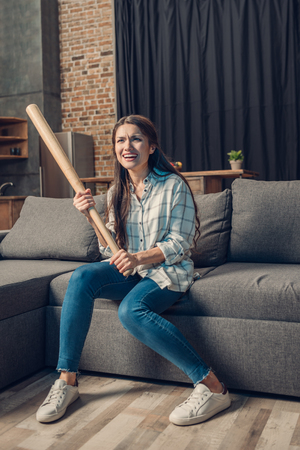 Young woman holding a baseball bat sitting on couch in living room and watching a game