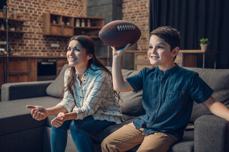 Cheering little boy and his mother sitting on couch with rugby ball while watching game on tvの写真素材